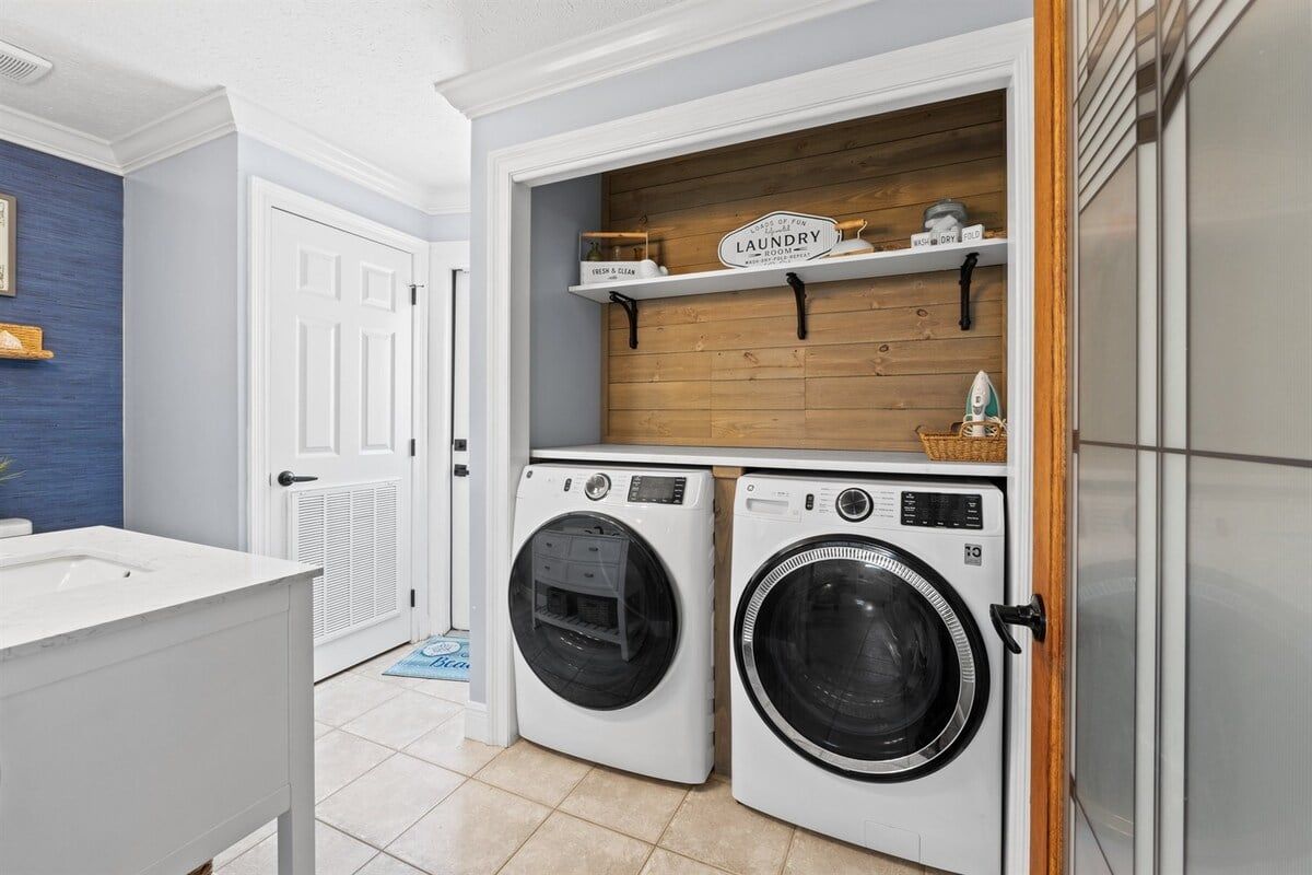 A laundry room with two front-loading washing machines, a wooden shelf above, and a white door to the left.