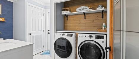 A laundry room with two front-loading washing machines, a wooden shelf above, and a white door to the left.