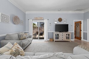 A living room with a grey sofa, a flat-screen TV on a wooden cabinet, and a sign reading 'BEACH HOUSE' above the door. - Hot Tub, Sleeps 10, 1 block to beach access, Pets (Panama City Beach)