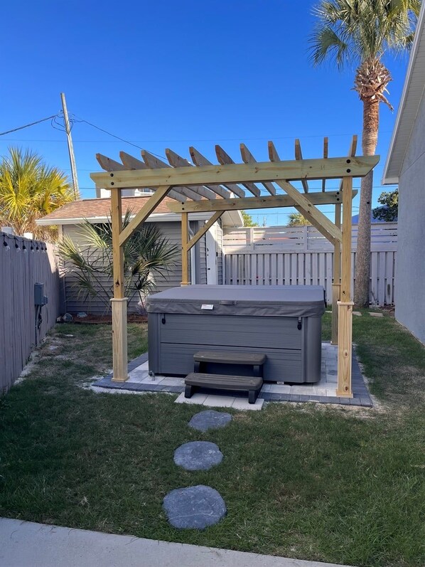 Un jacuzzi bajo una pérgola de madera en un patio trasero con una palmera y una valla blanca.