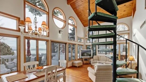 A spacious living area with a wooden floor, a staircase, and large windows offering a view of the outdoors. - Luxury Meets the Lake (Lake George)