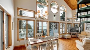 A spacious living room with a wooden floor, a dining area with white chairs, a sofa, and a TV. Large windows provide natural light and views of the outdoors. - Luxury Meets the Lake (Lake George)