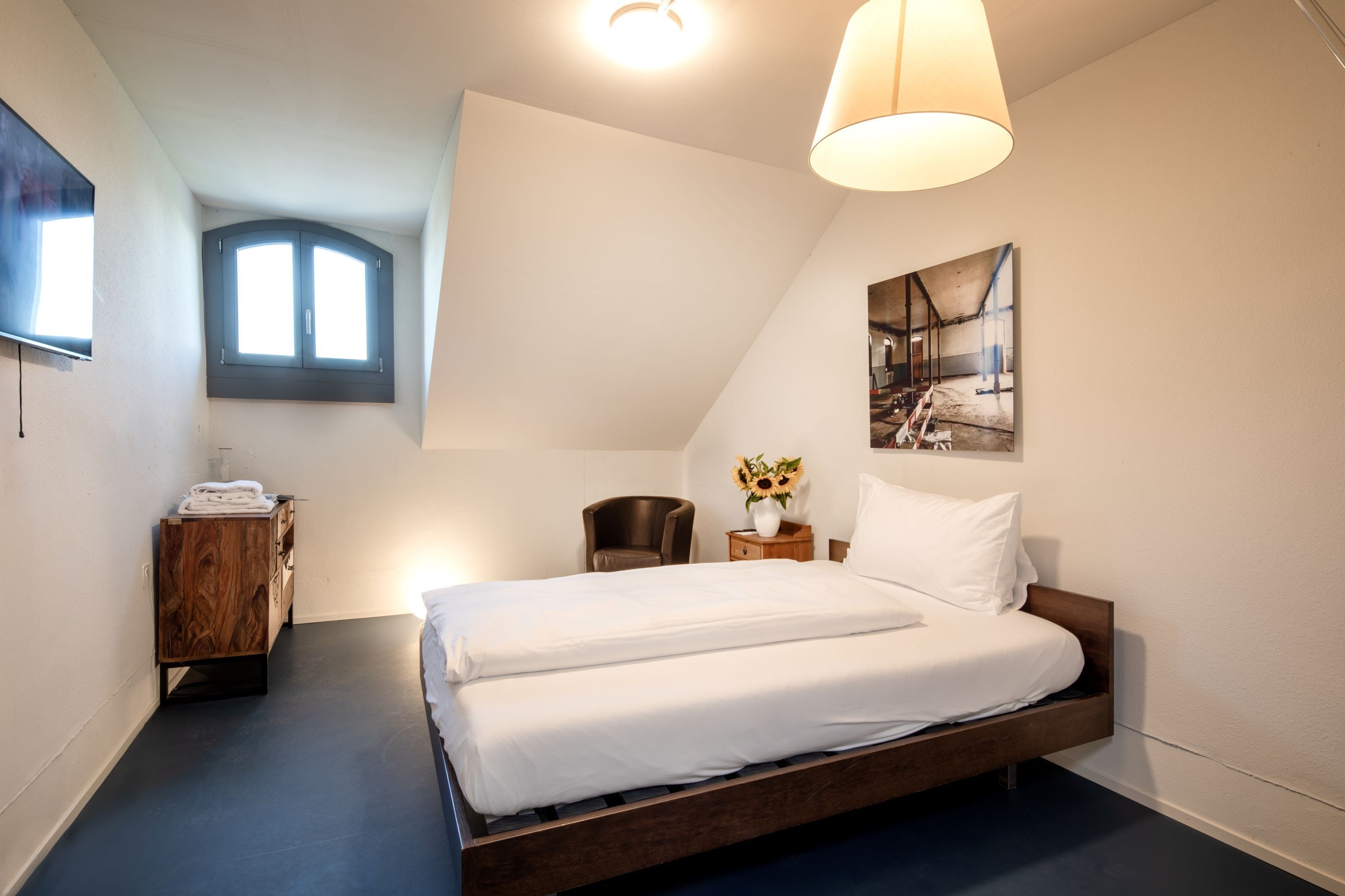 Un dormitorio con una cama grande, una mesita de noche de madera, una silla, un ventanal y un cuadro en la pared.