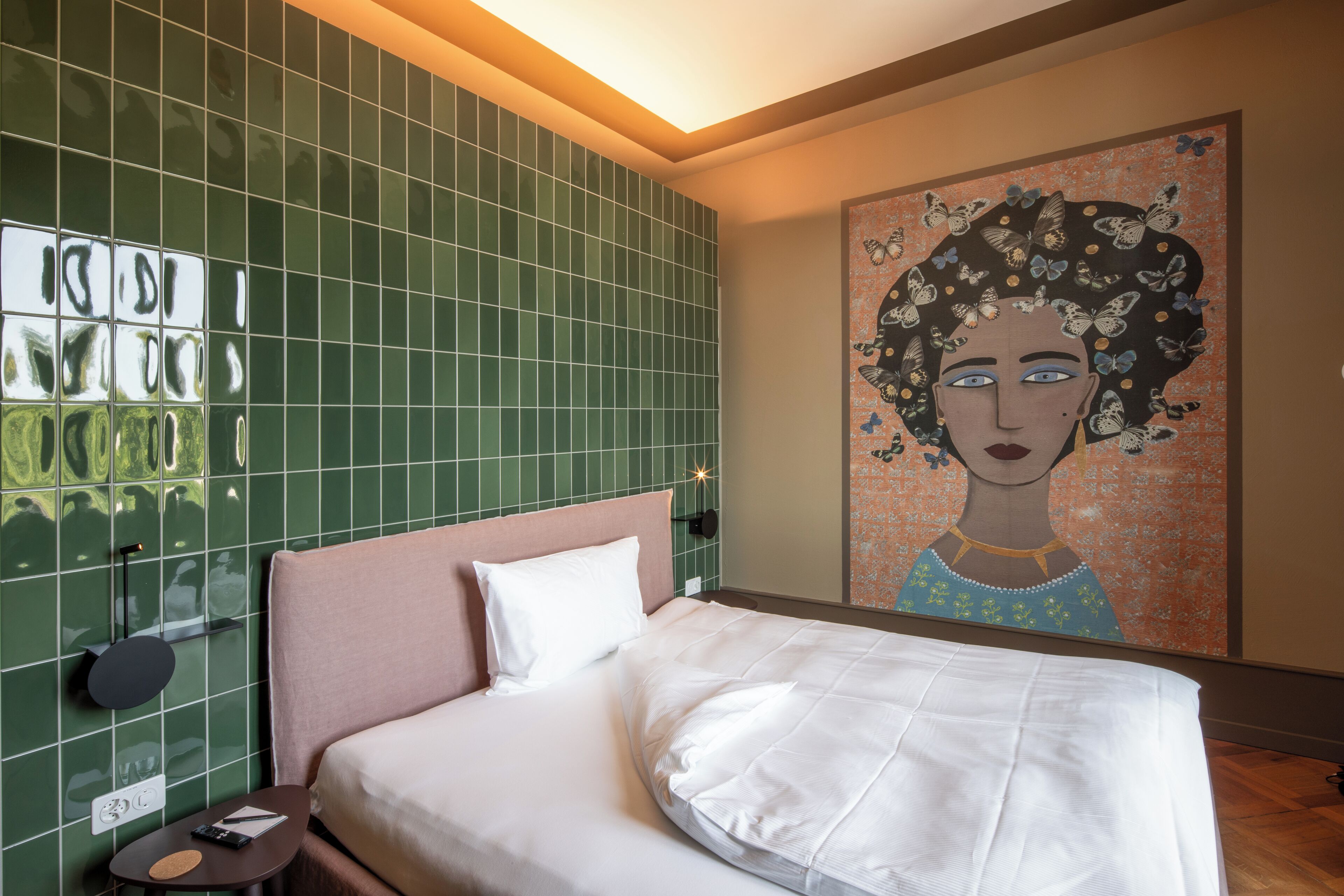 Una habitación de hotel con cama, mesita de noche, pared de azulejos verdes y un cuadro.