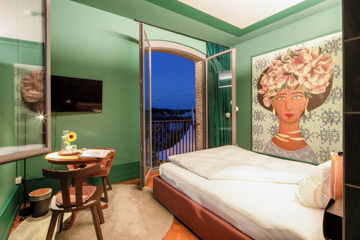 Ein Schlafzimmer mit Bett, Sessel, Tisch mit Vase und Blume, einem Wandbild und einem Balkon mit Blick auf die beleuchtete Stadt.