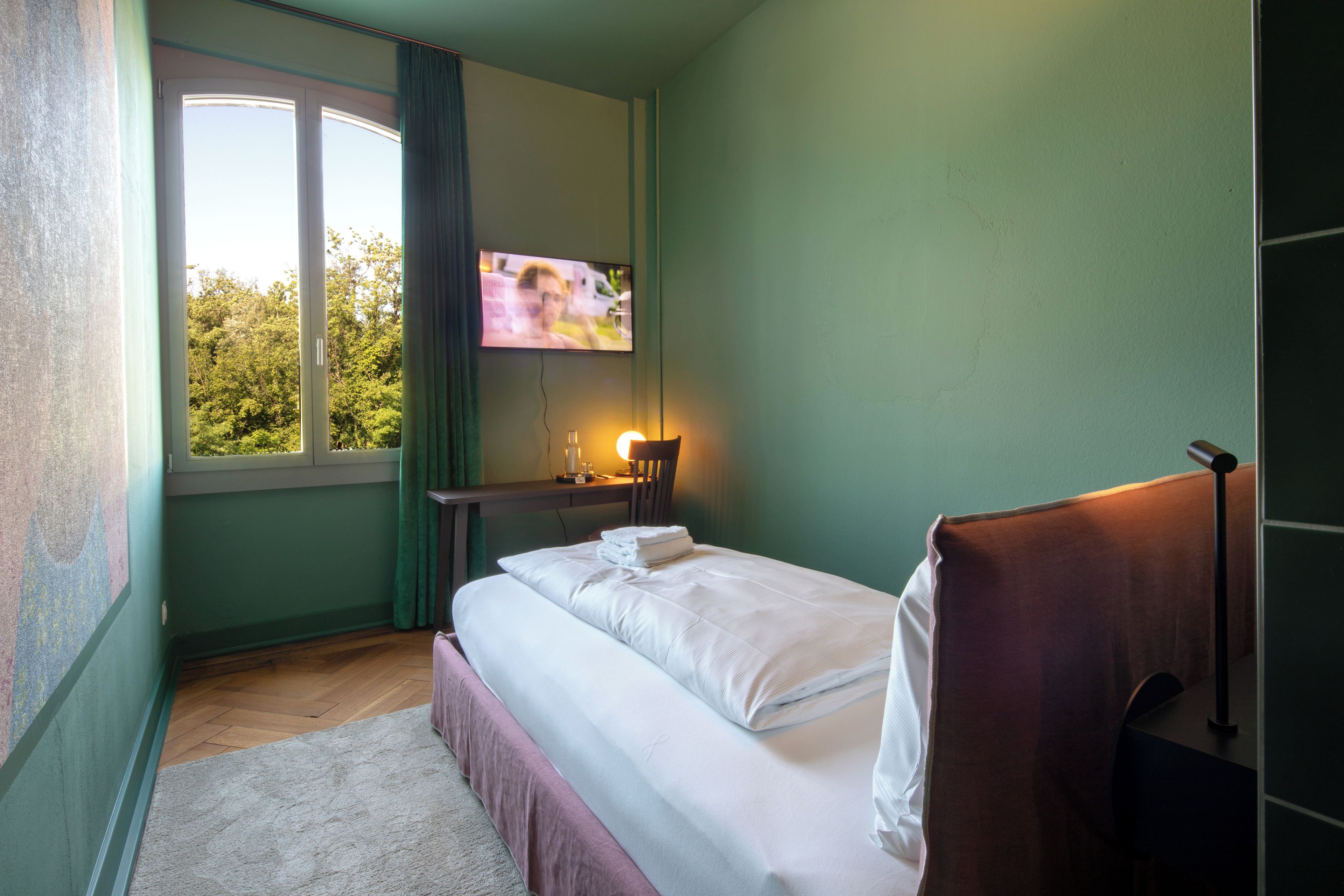 A hotel room with a bed, a TV mounted on the wall, a bedside table with a lamp, and a window with curtains.