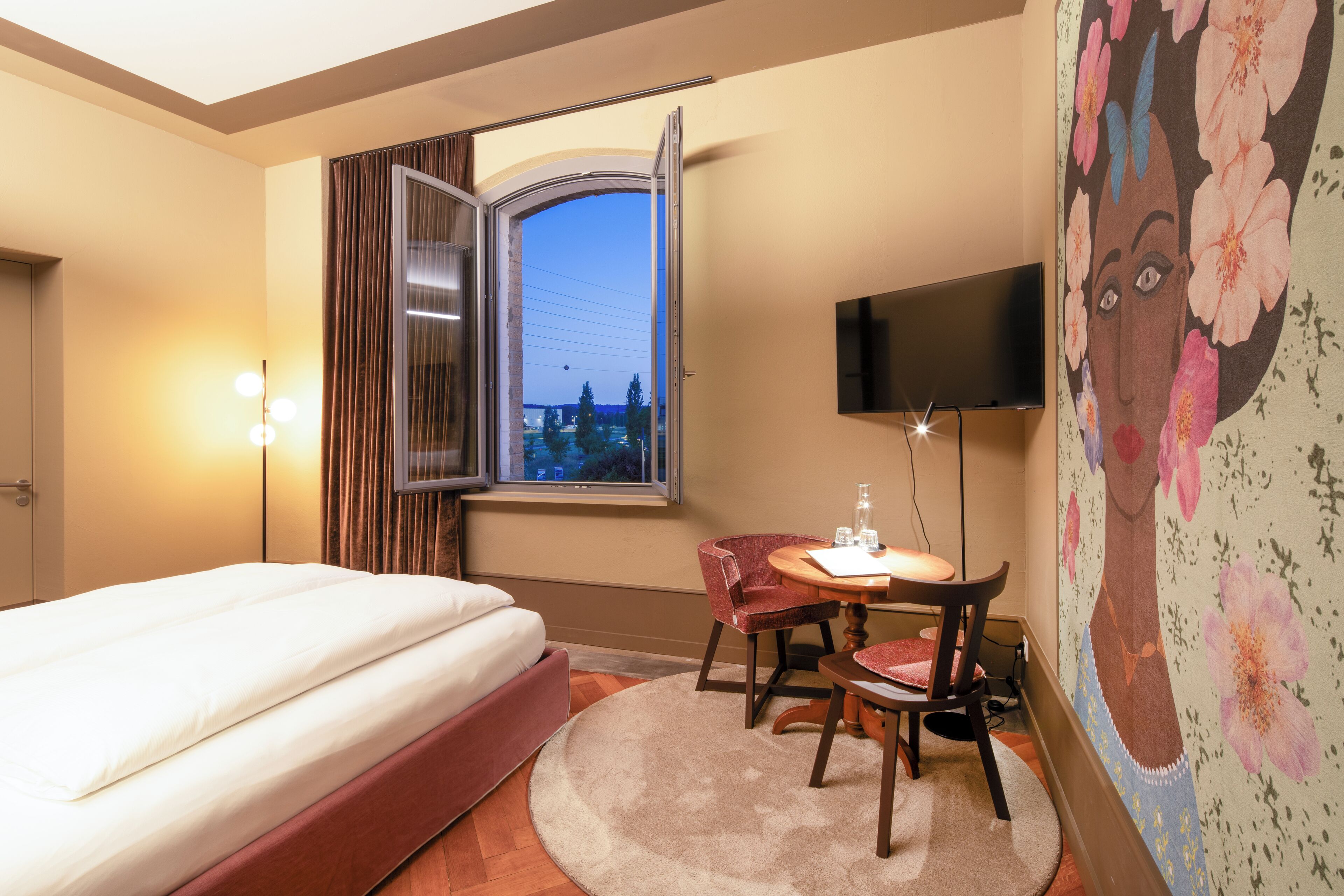 A hotel room with a bed, a desk with a chair, a television, and a window with curtains.