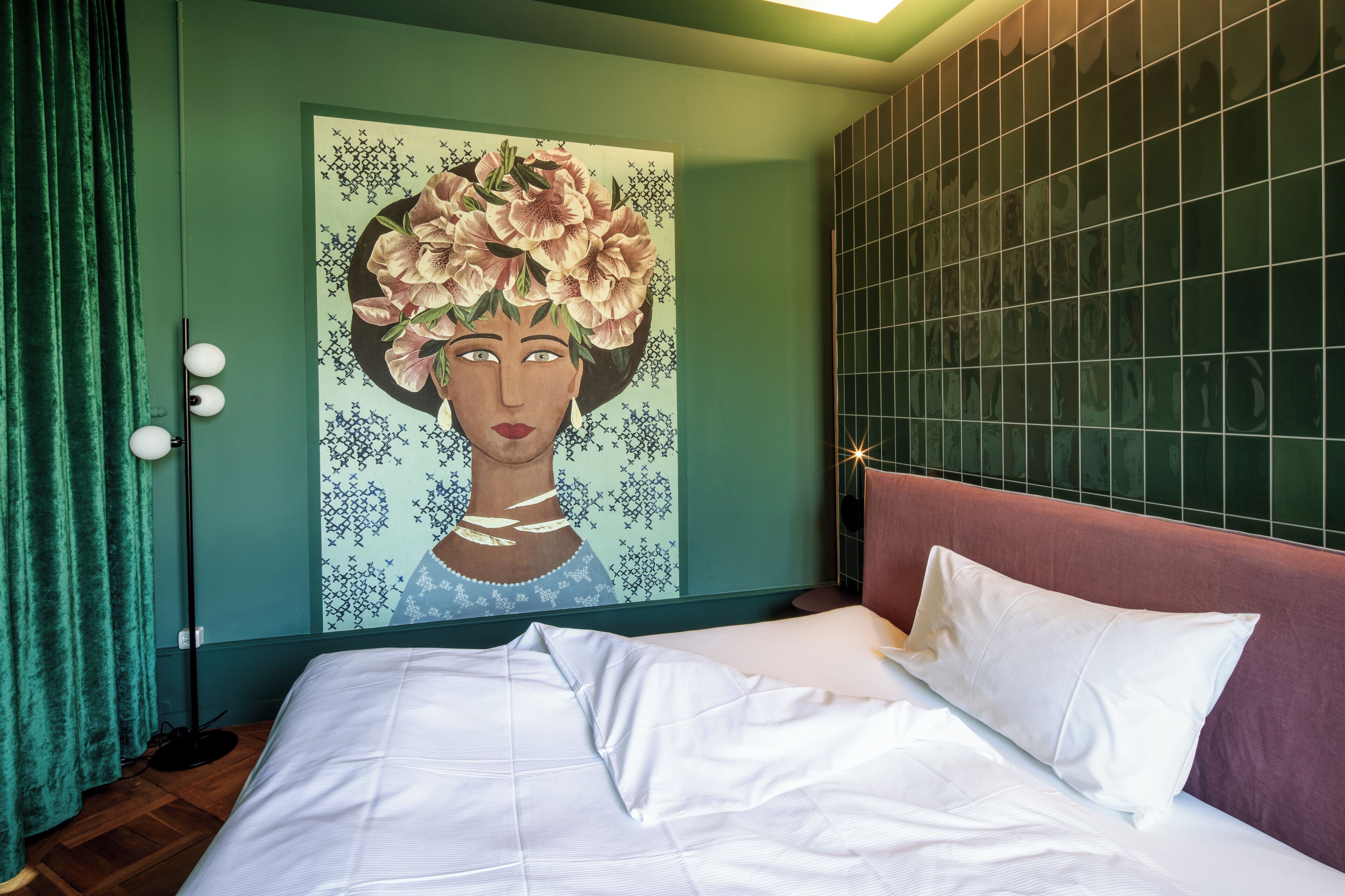 Un dormitorio con cama, cortinas verdes, una lámpara de pie y un cuadro en la pared.