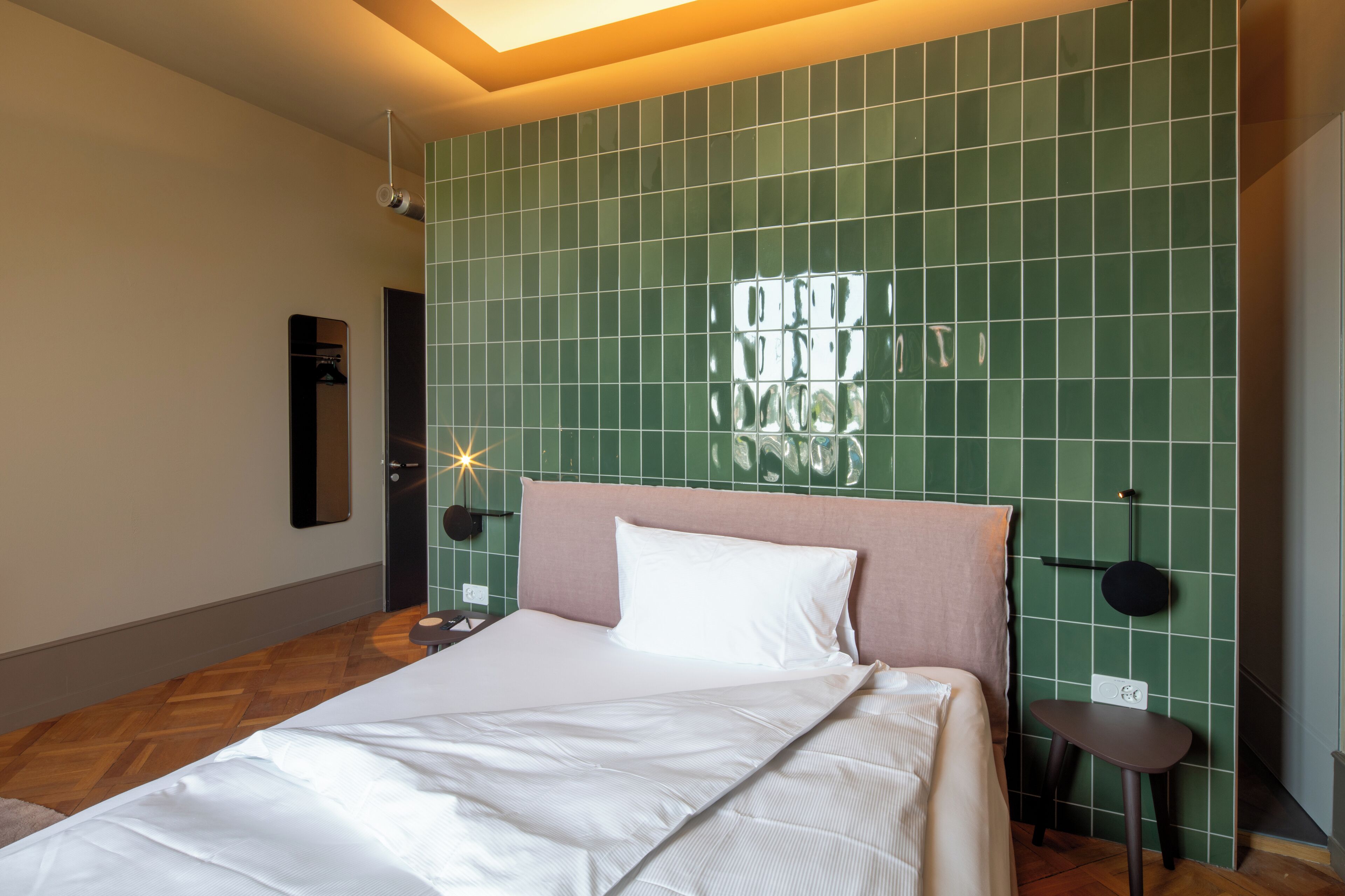 A neatly made bed with white linens, a green tiled wall, and a small bedside table with a lamp.