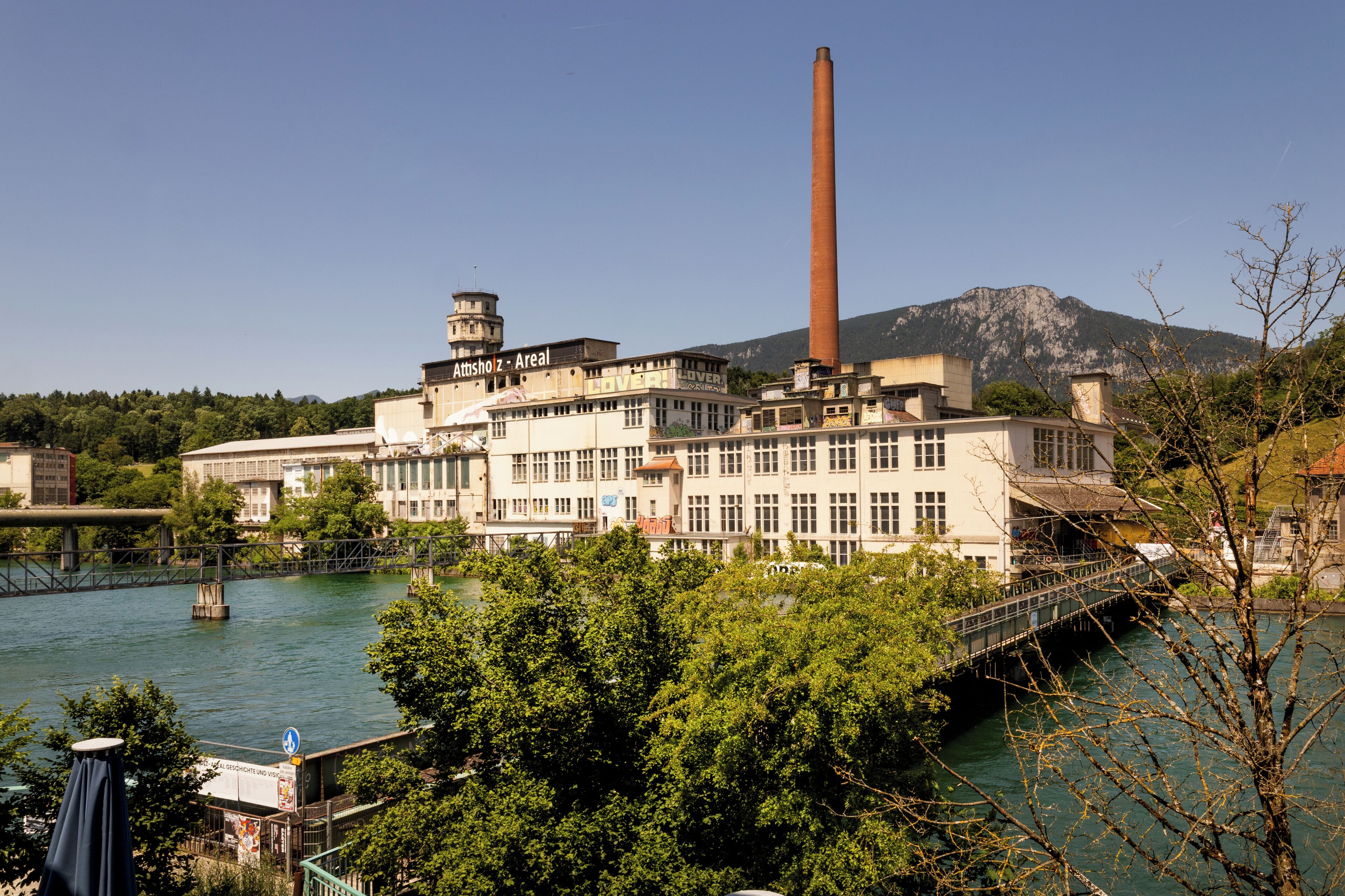 A large industrial building with a tall chimney, a bridge over a river, and a mountainous backdrop.
