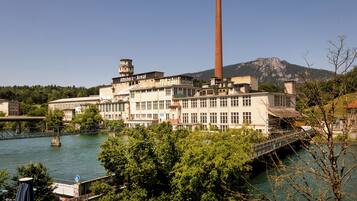 A large industrial building with a tall chimney, a bridge over a river, and a mountainous backdrop.