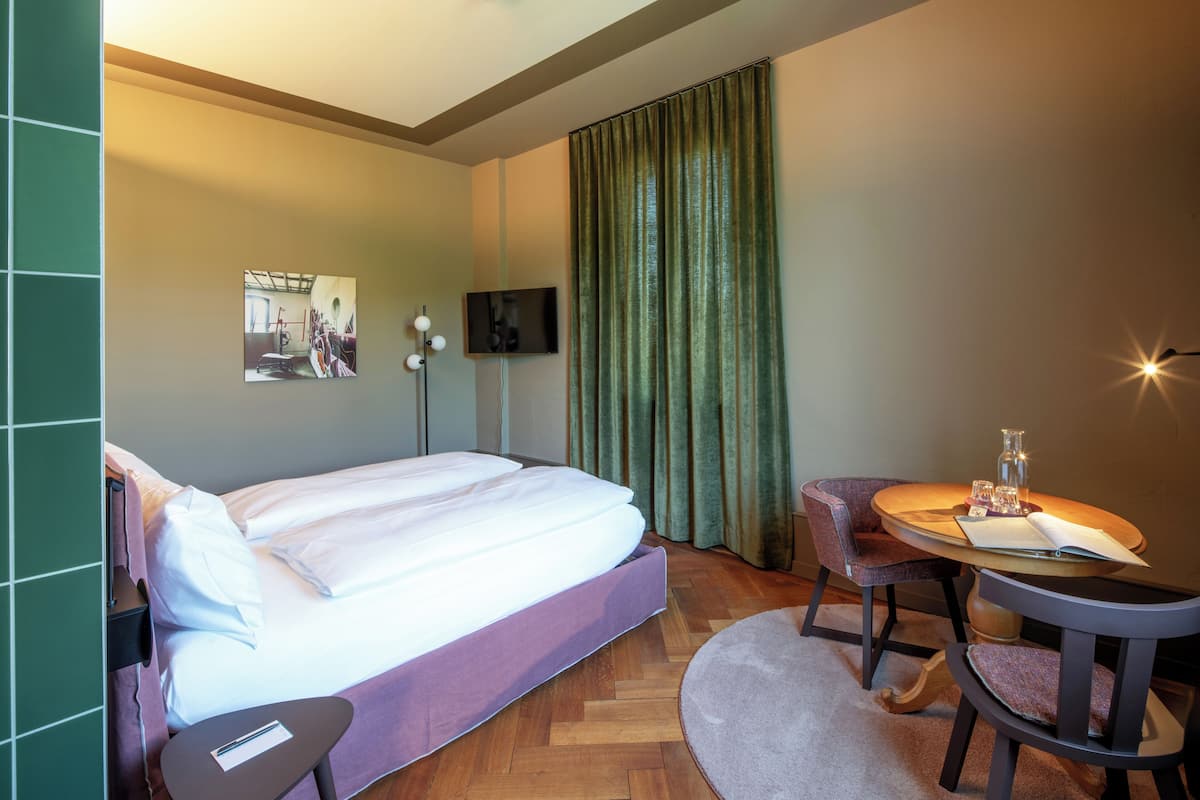 Ein Hotelzimmer mit einem Bett, einem kleinen Tisch mit Stuhl, einem Fernseher und einem Fenster mit Vorhängen.