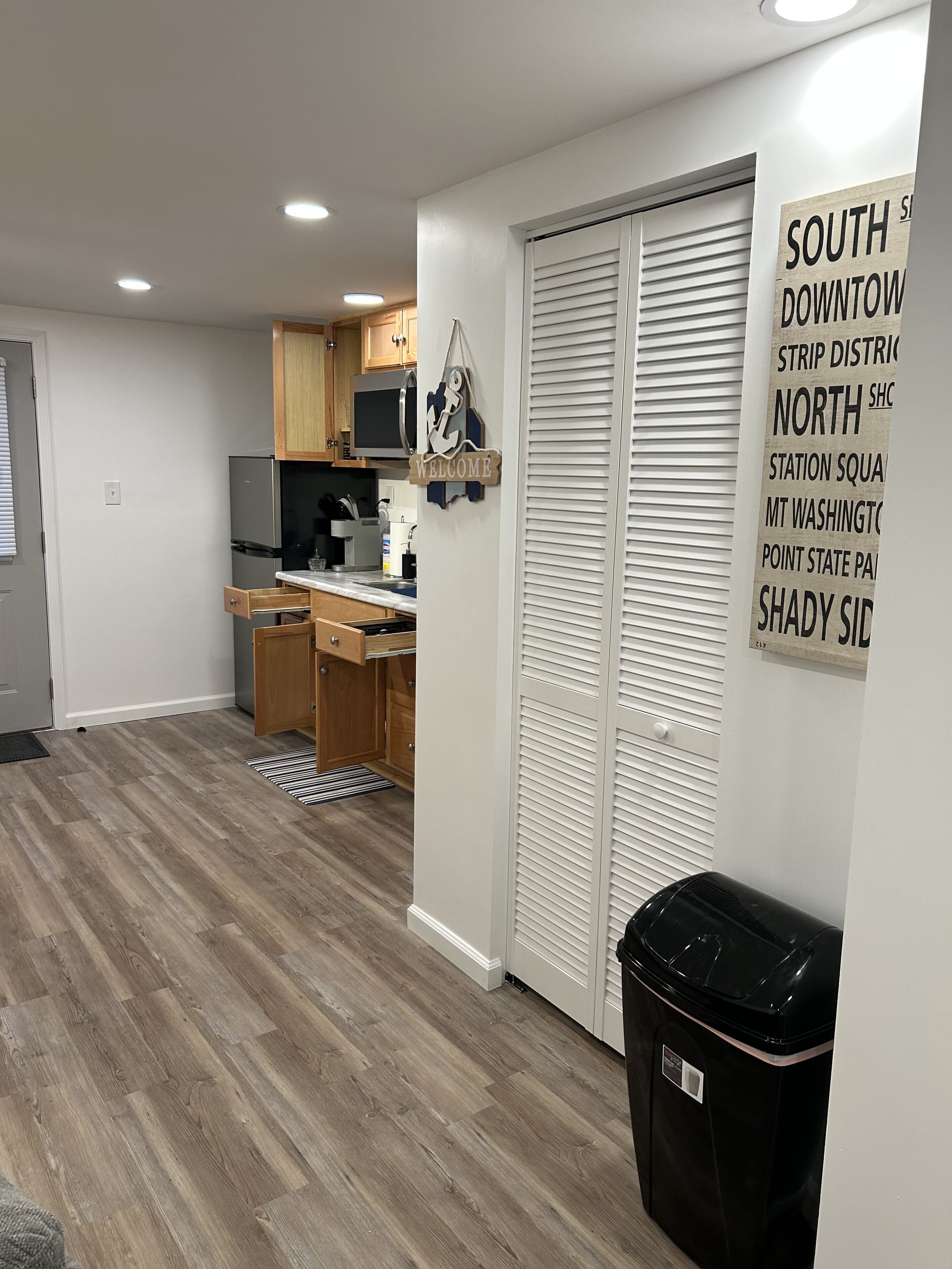 A modern kitchen with wooden cabinets, a stainless steel refrigerator, and a microwave. There is a black trash bin and a welcome sign on the wall.