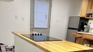 A kitchen with a wooden countertop, two stools, a window with blinds, and a refrigerator.