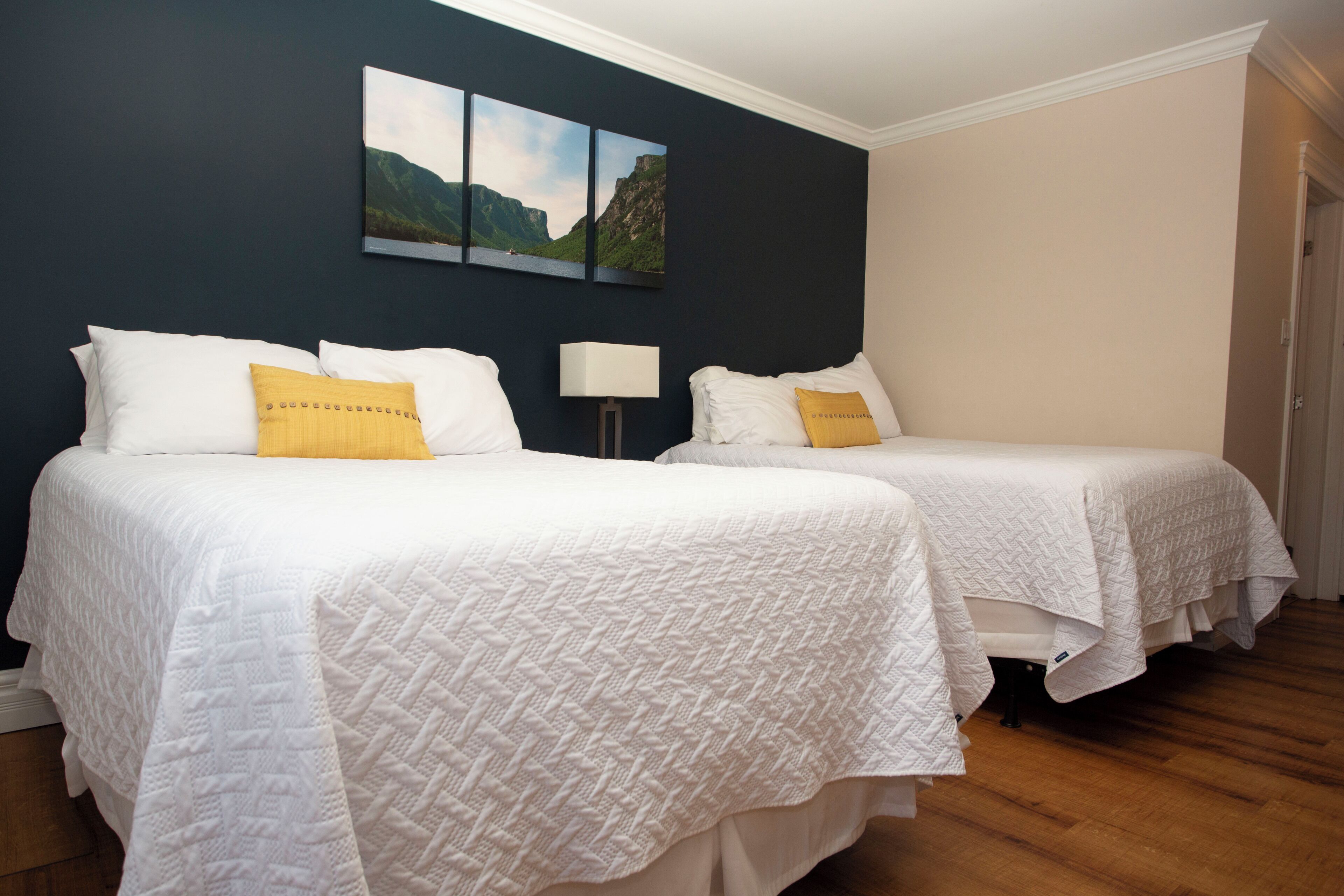 A hotel room with two beds, each with white bedding and a yellow accent pillow. There is a painting above the beds and a lamp between them.