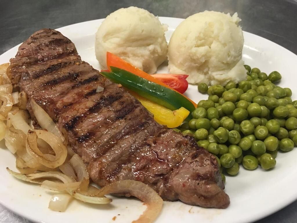 A plate with grilled steak, mashed potatoes, sautéed onions, bell peppers, peas, and a slice of tomato.