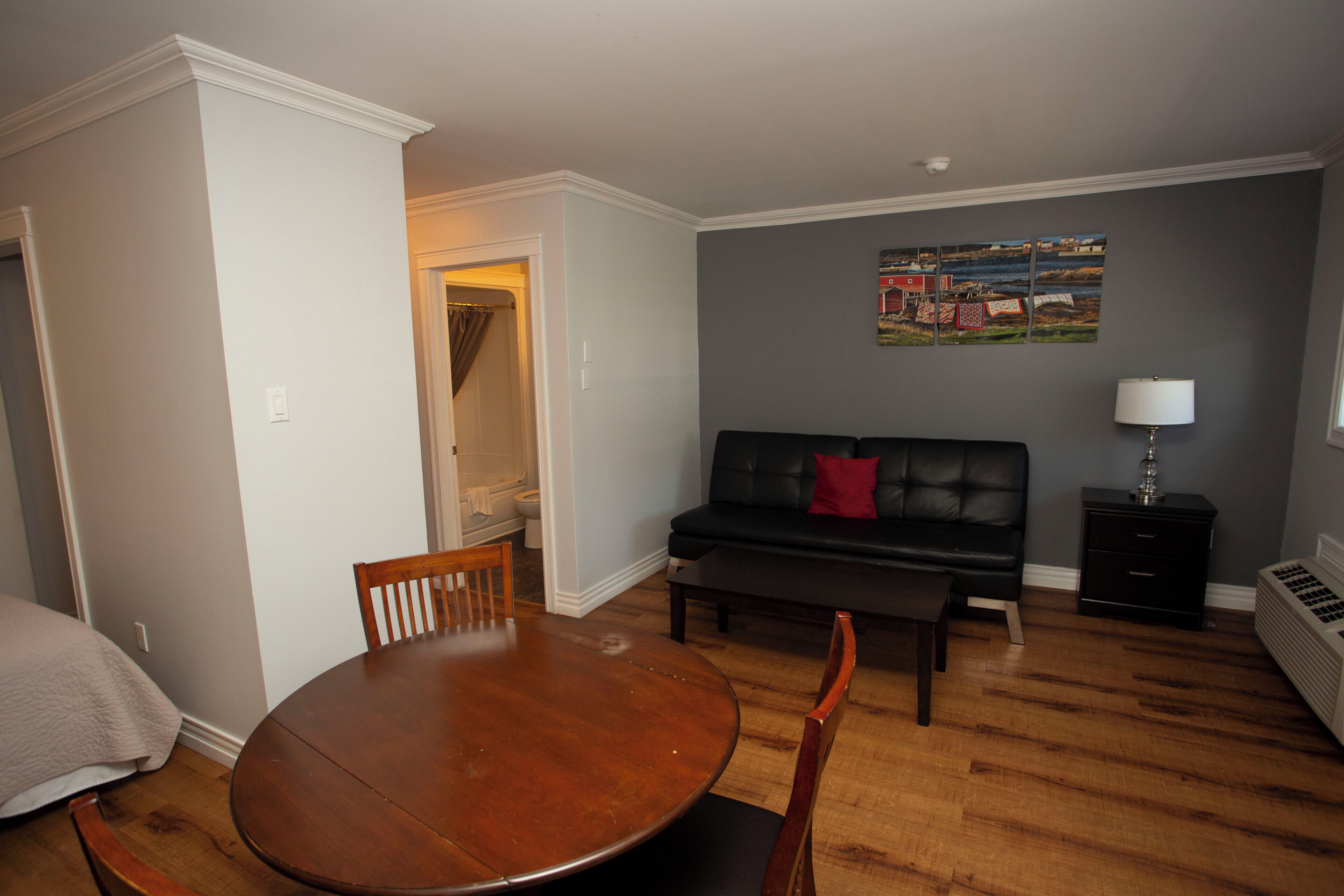 A living room with a round wooden table, two wooden chairs, a black sofa with a red pillow, a black cabinet, and a wall-mounted lamp.