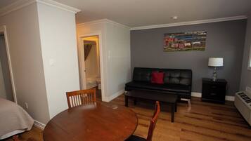 A living room with a round wooden table, two wooden chairs, a black sofa with a red pillow, a black cabinet, and a wall-mounted lamp.