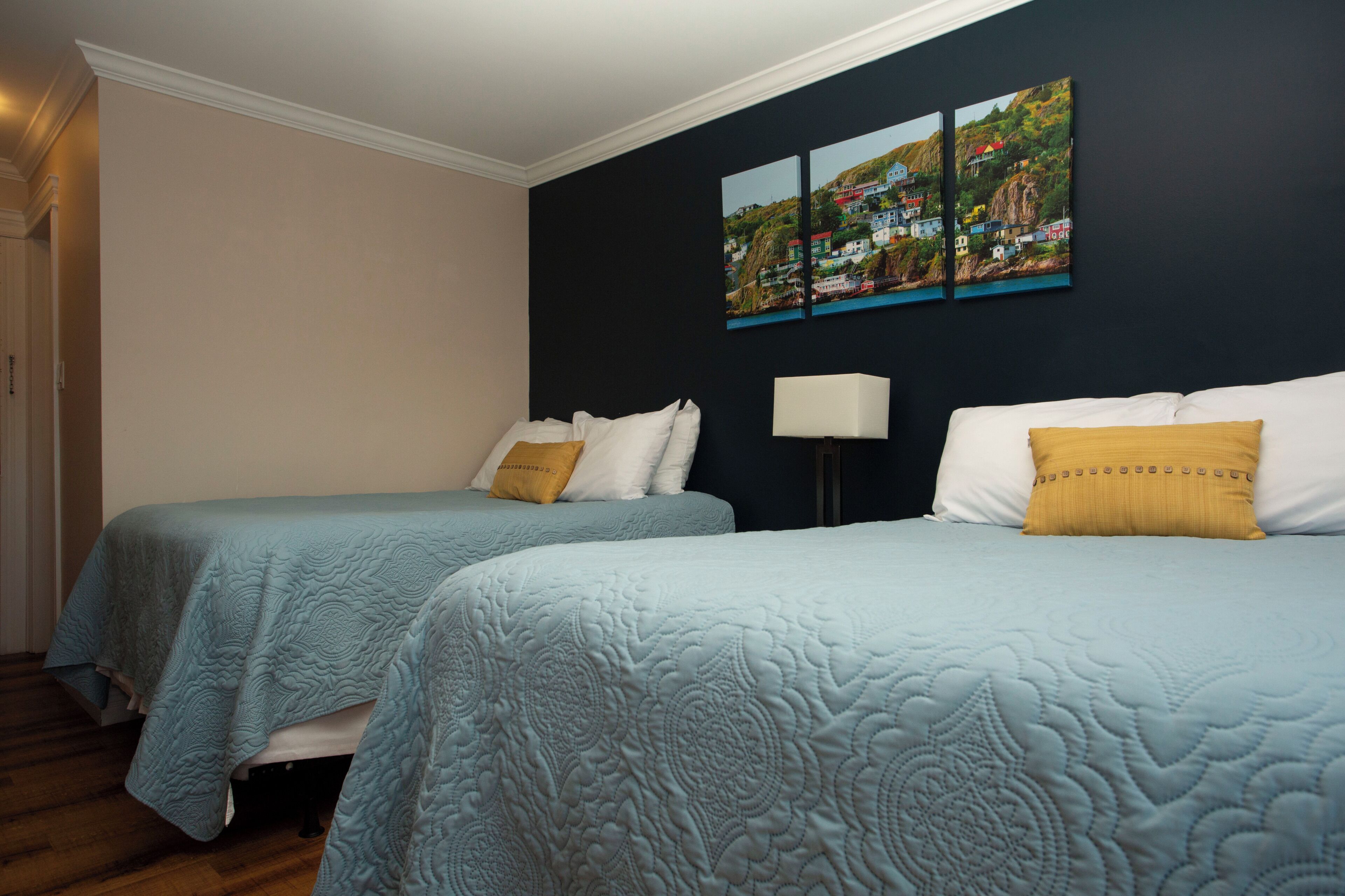 A hotel room with two beds, each with a blue quilted bedspread and matching pillows, a bedside lamp, and a wall-mounted painting of a coastal town.