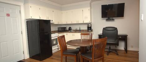 Cocina compacta con armarios blancos, un refrigerador negro, microondas y cafetera. Hay una mesa de comedor con sillas y una silla de oficina negra. Un televisor de pantalla plana está montado en la pared.