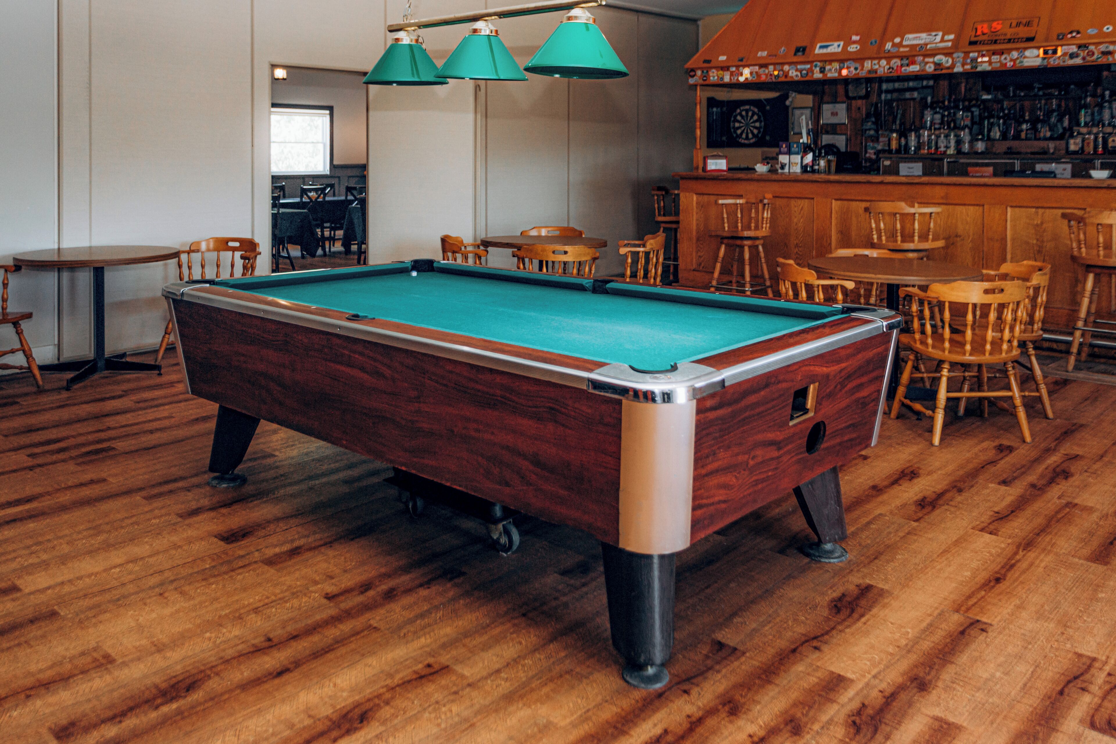 Una mesa de billar en una sala con piso de madera, rodeada de sillas y una zona de bar con taburetes.