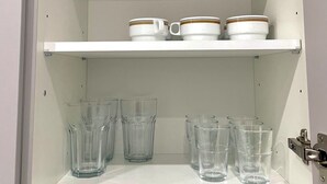 A white cabinet with two shelves; the top shelf holds two white mugs with gold trim, and the bottom shelf contains six clear glass tumblers. - Small and fine city apartment in Saarbrücken (Saarbrücken)