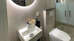 A modern bathroom with a round mirror, a white sink, a toilet, and a glass-enclosed shower. - Small and fine city apartment in Saarbrücken (Saarbrücken)