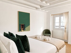 A hotel room with a bed, pillows, a chair, a window with curtains, and a painting on the wall.