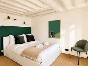 A neatly made bed with white linens and a green headboard, a single green chair, and a framed picture on the wall.