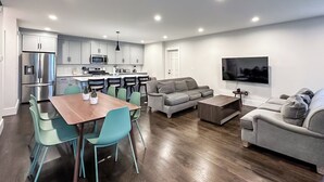 Una cocina moderna con electrodomésticos de acero inoxidable, una zona de comedor con una mesa de madera y sillas color turquesa, un sofá gris y un televisor de pantalla plana montado en la pared.