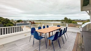 Un porche cubierto con una mesa de madera y sillas azules, con vistas a una masa de agua y edificios lejanos.