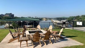 A patio with wooden chairs and tables overlooking a body of water, with a view of buildings and a bridge in the distance. - Oasis Luxury Lakefront Condo:1st Floor, Pool, HotTub, Dining, Boating, Fishing! (Graford)