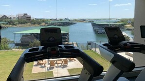 A view from a room with exercise equipment, overlooking a body of water with docks and buildings in the background. - Oasis Luxury Lakefront Condo:1st Floor, Pool, HotTub, Dining, Boating, Fishing! (Graford)