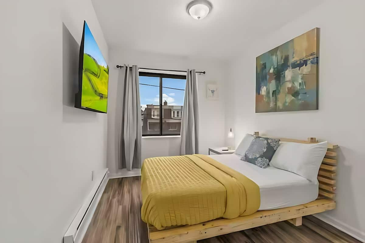A bedroom with a bed, a TV, a window with curtains, and a painting on the wall.
