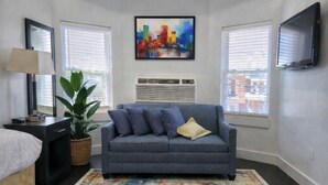 Un dormitorio moderno con un sofá gris, una alfombra colorida, una planta grande y una pintura de un paisaje urbano en la pared.