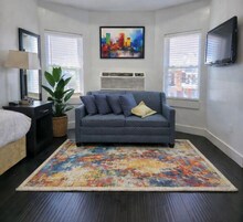 Un dormitorio moderno con un sofá gris, una alfombra colorida, una planta grande y una pintura de un paisaje urbano en la pared.