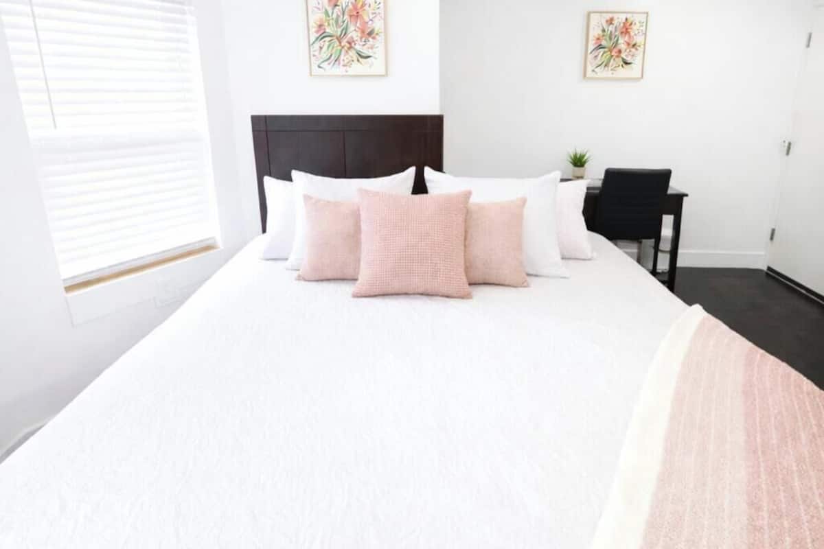 A neatly made bed with white and pink bedding, a dark headboard, and two floral paintings on the wall.