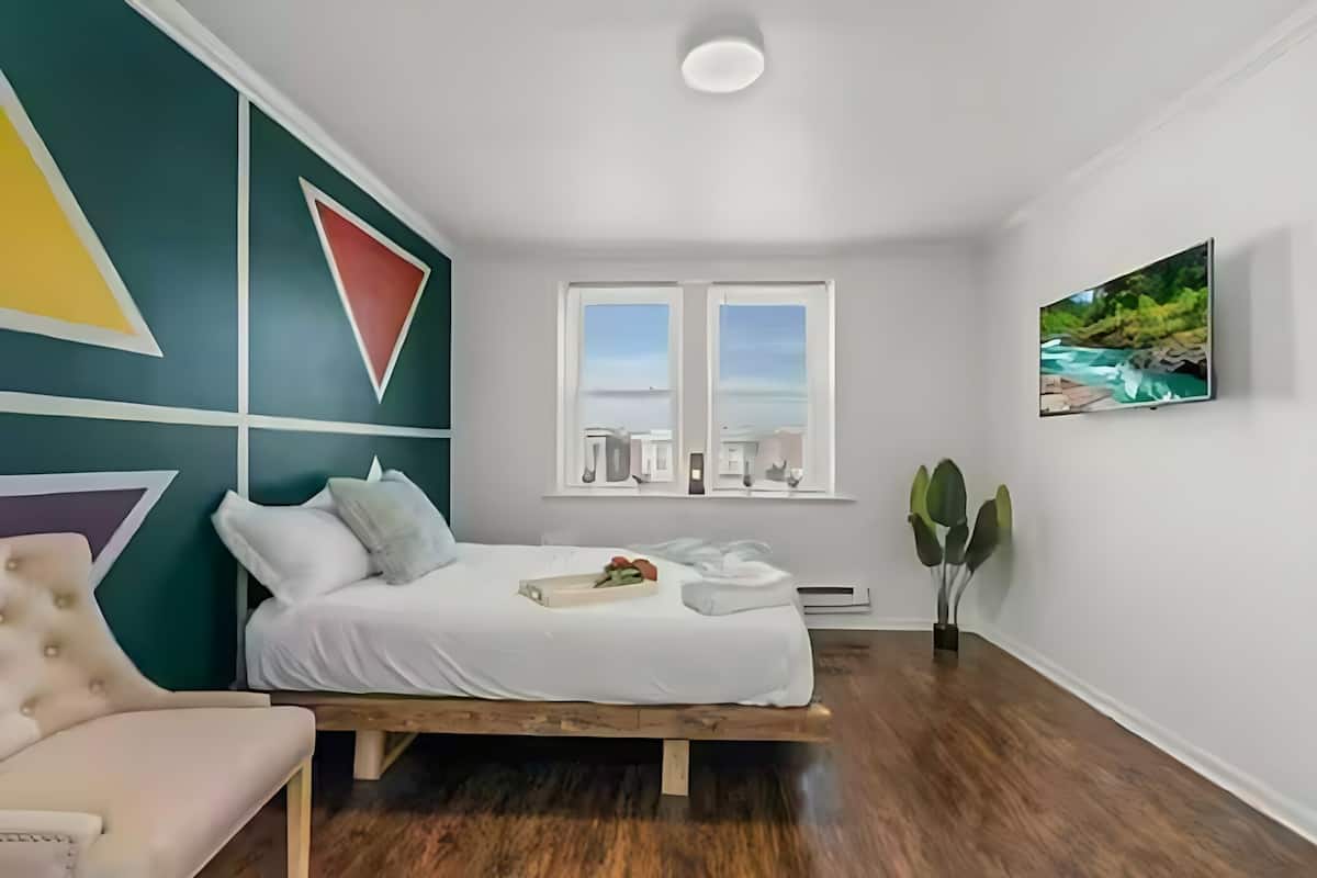 A bedroom with a bed, a chair, a window with a view, and a painting on the wall.