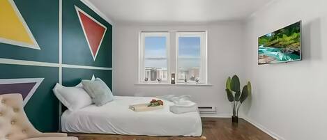 A bedroom with a bed, a chair, a window with a view, and a painting on the wall.