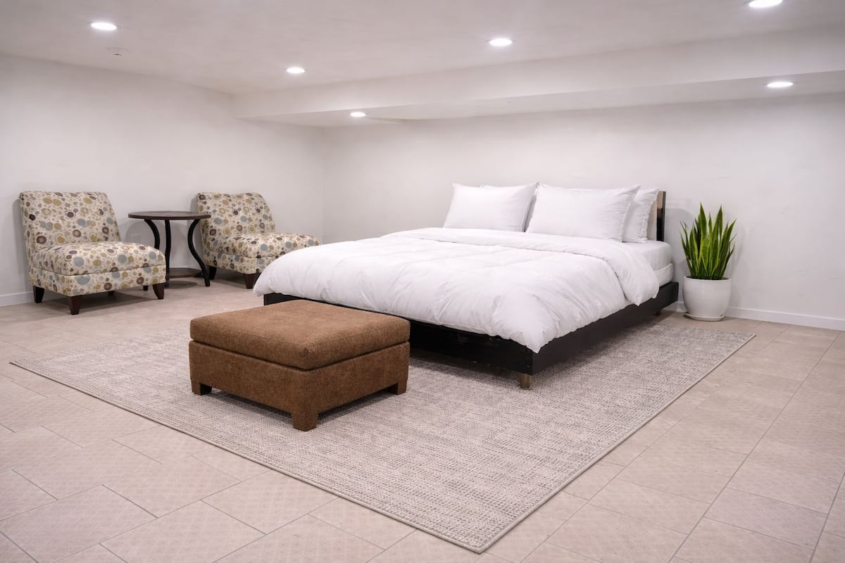 A bedroom with a bed, two armchairs, a small table, a brown ottoman, and a potted plant.