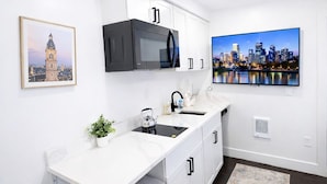 Una cocina moderna con muebles blancos, un microondas integrado, un lavamanos y un cuadro de un paisaje urbano en la pared.