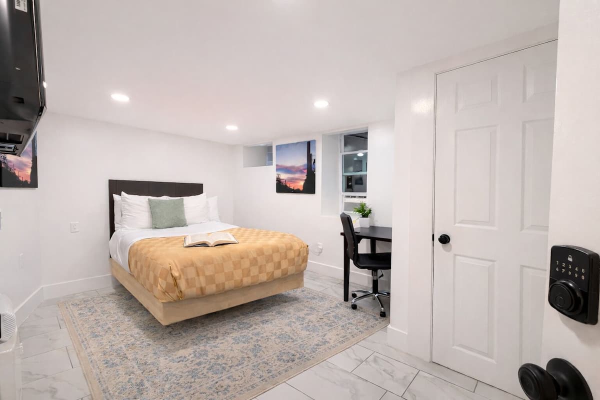 A hotel room with a bed, a desk, a chair, a door with a keypad lock, and a small window with a cityscape view.