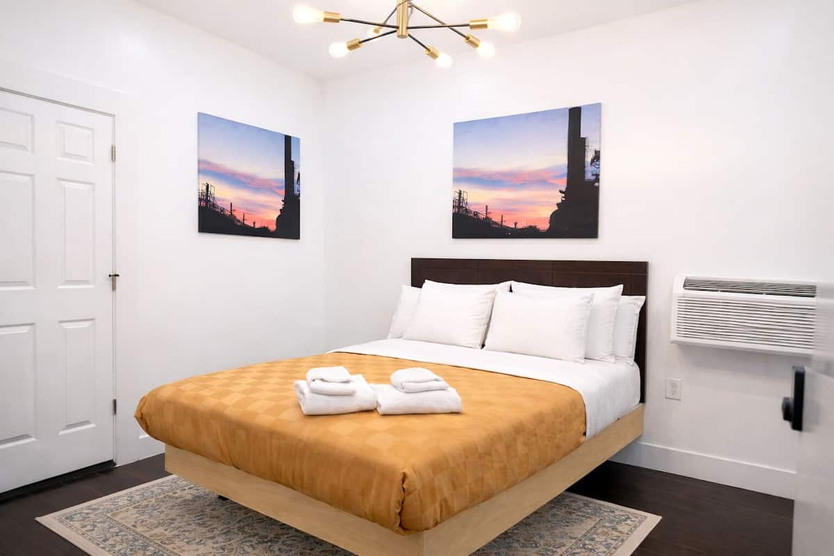 A hotel room with a bed, white pillows, a brown bedspread, two wall-mounted paintings, a ceiling light fixture, and a wall-mounted air conditioner.