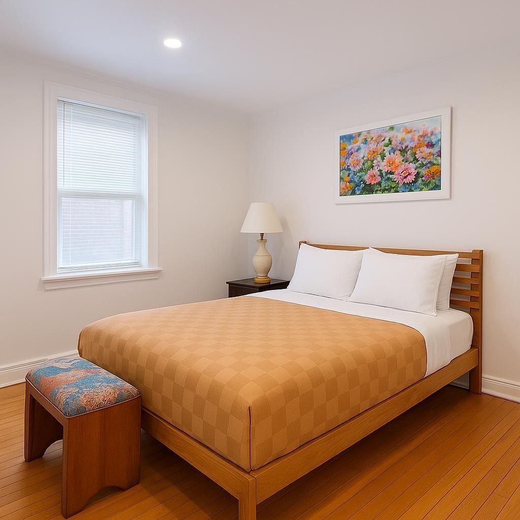 A bedroom with a wooden bed, a nightstand with a lamp, a window with blinds, and a floral painting on the wall.