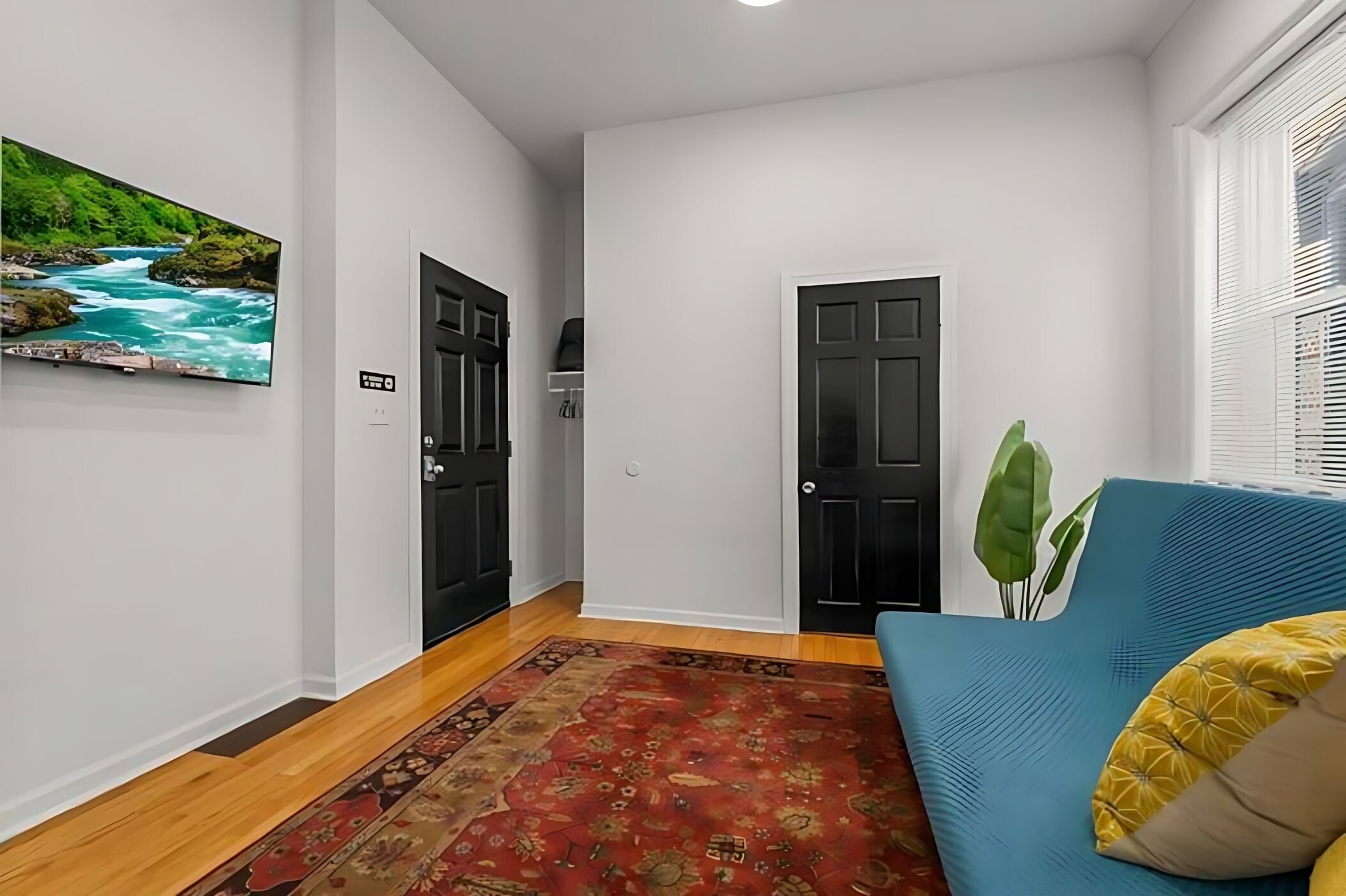 A hallway with a red patterned rug, a blue sofa with yellow cushions, a black door, and a wall-mounted TV displaying a river scene.