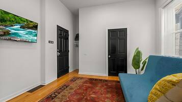 A hallway with a red patterned rug, a blue sofa with yellow cushions, a black door, and a wall-mounted TV displaying a river scene.