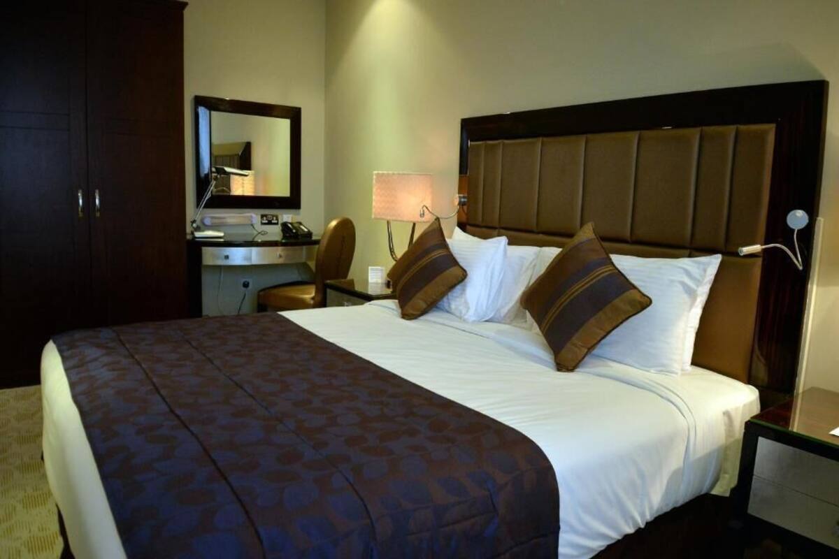 A hotel room with a large bed, a desk with a lamp, a chair, and a wardrobe.
