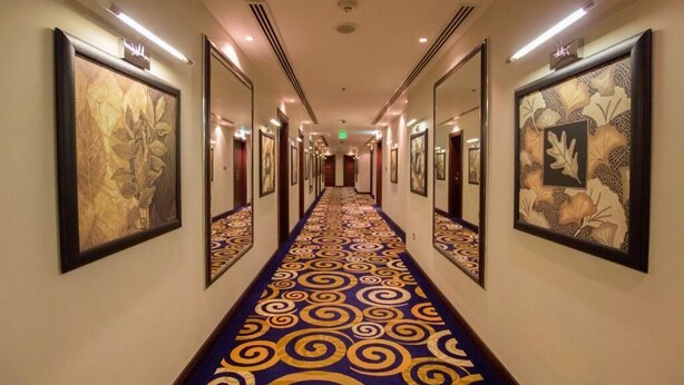 A hotel hallway with a patterned carpet, framed artwork on the walls, and a long corridor leading to an exit. - Premium Strato Hotel (Doha)