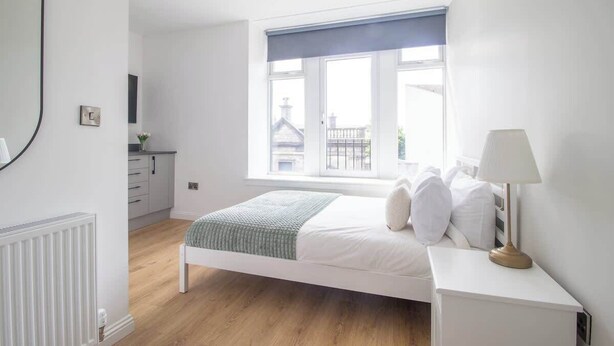A bedroom with a bed, bedside table, lamp, window, and a view of buildings outside. - Double Ensuite Room + Shared Kitchen + Fast Wi-Fi (Grangemouth)