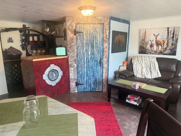 Una sala de estar acogedora, con una puerta roja y azul, un sofá de cuero marrón, una mesa de comedor con vasos transparentes y un cuadro de ciervos en la pared.